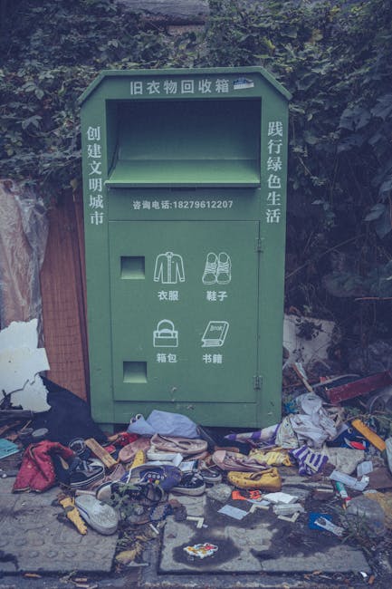 An outdoor scene showing a collection of discarded environmental waste, primarily consisting of multiple stacks of flattened cardboard fruit and vegetable packaging boxes with vibrant printed labels, 