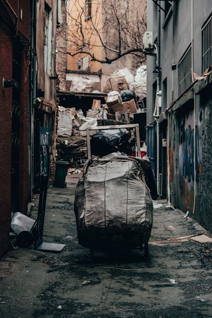 A narrow urban alleyway cluttered with a large, overfilled grey garbage bag in the foreground, atop a paved surface that shows signs of dirt and minor debris. Behind the bag, a collection of irregular