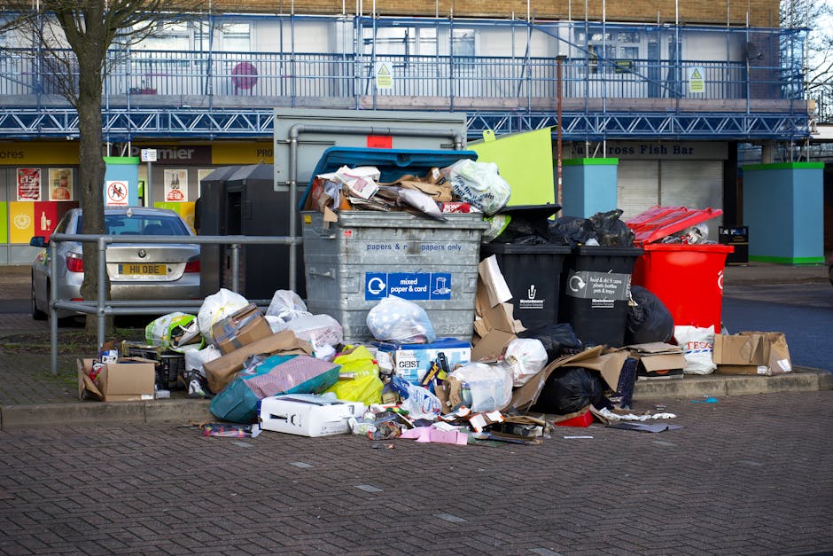A narrow urban alleyway cluttered with a large, overfilled grey garbage bag in the foreground, atop a paved surface that shows signs of dirt and minor debris. Behind the bag, a collection of irregular